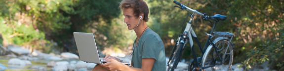 Mann sitzt mit einem Computer an einem steinigen Flussufer